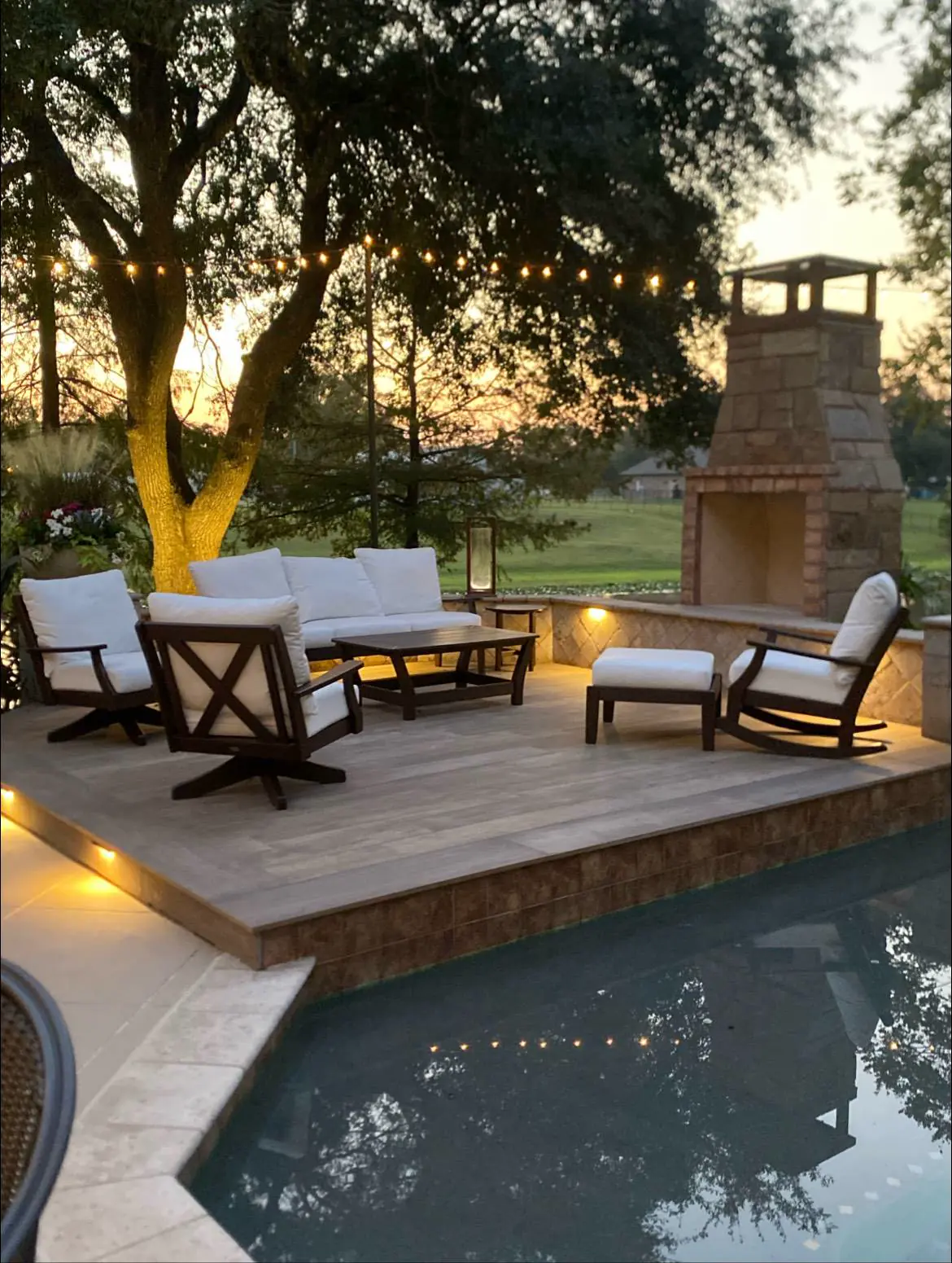 Stone outdoor fireplace on a wood deck with cushioned seating, string lights, and a pool at dusk